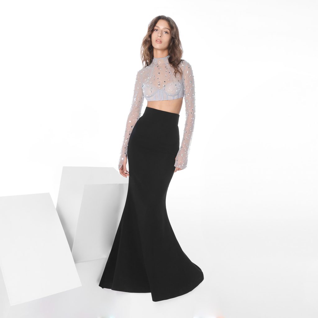 A high‑neck, corseted long‑sleeve cropped top embellished with confetti crystals and beads paired with a mermaid‑cut long skirt, with the full look also available as a single dress.